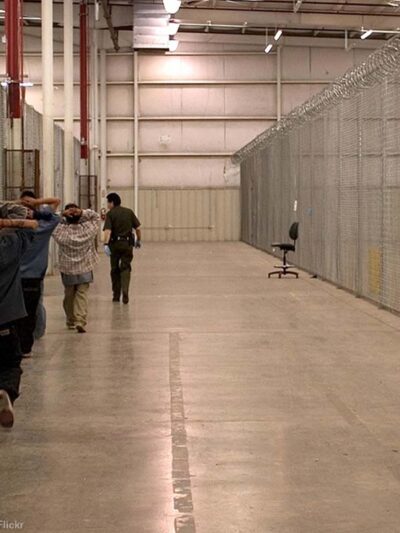 Immigrant detainees walk down a hall escorted by heavily armed CBP officers.