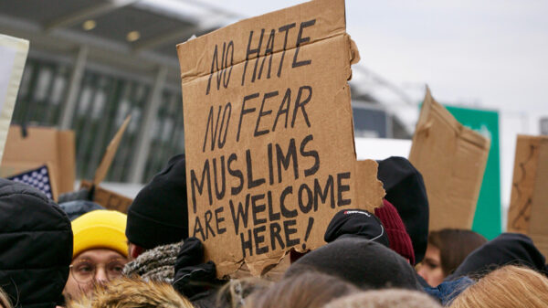 Person holding cardboard sign reading "No Hate / No Fear / Muslims Are Welcome Here!" at a protest at an airport in opposition to the Trump "Muslim Ban"