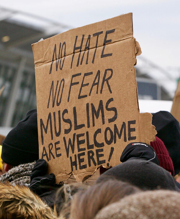 Person holding cardboard sign reading "No Hate / No Fear / Muslims Are Welcome Here!" at a protest at an airport in opposition to the Trump "Muslim Ban"