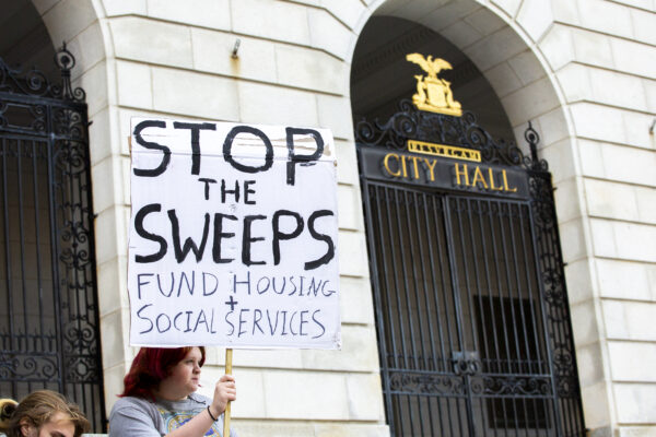 Person holding poster reading "stop the sweeps / fund housing + social services" in front of Portland City Hall.