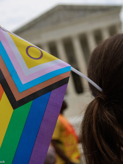 A Pride supporter (whose face cannot be seen) holds an Intersex-inclusive Pride Flag in their hair while facing the Supreme Court.