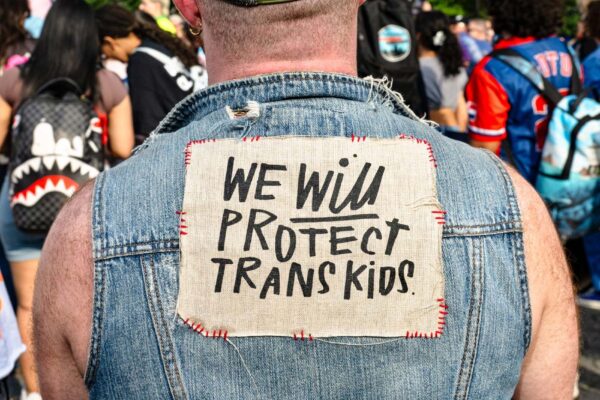At a trans rally, person (wearing a denim vest and with their back to the camera) displays the sign stitched to the vest that reads "WE WILL PROTECT TRANS KIDS."