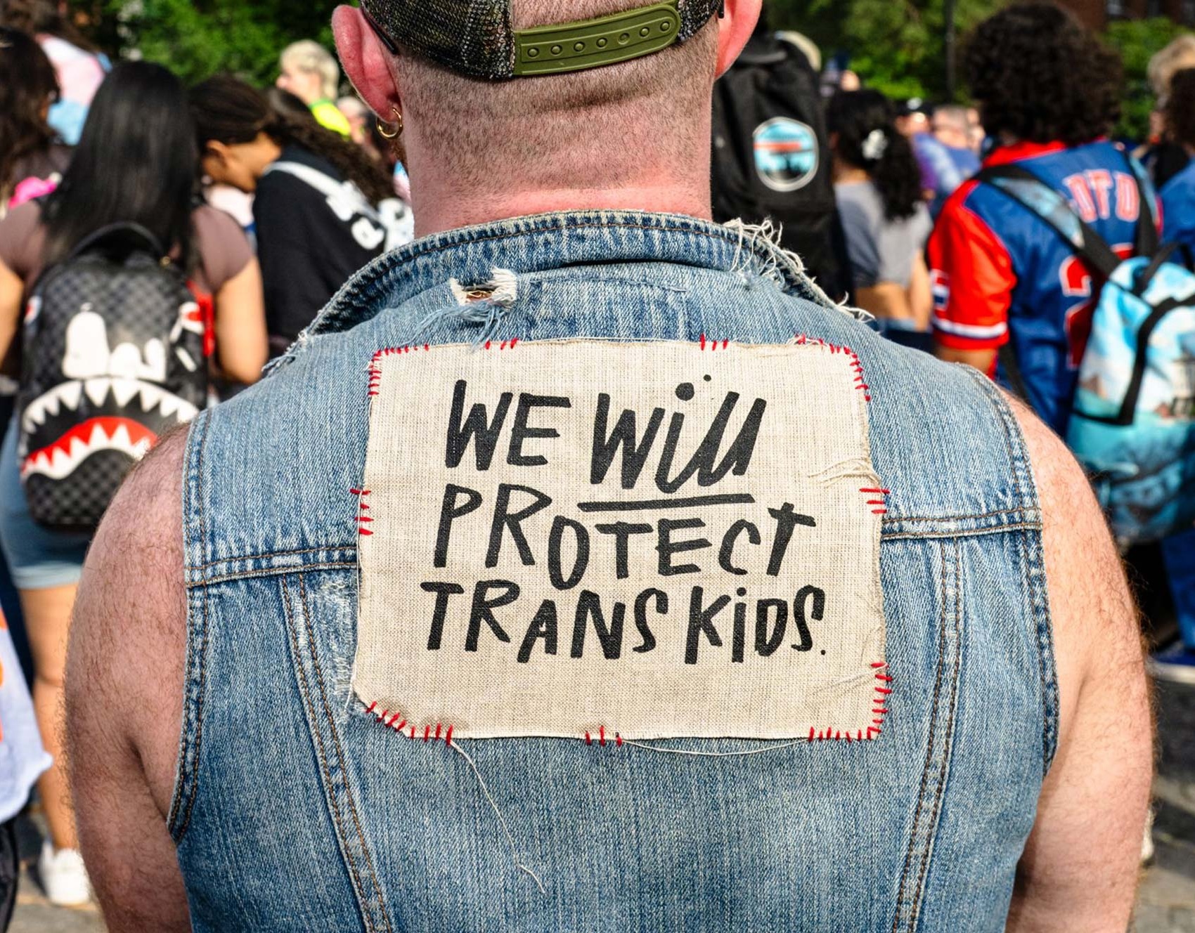 At a trans rally, person (wearing a denim vest and with their back to the camera) displays the sign stitched to the vest that reads "WE WILL PROTECT TRANS KIDS."