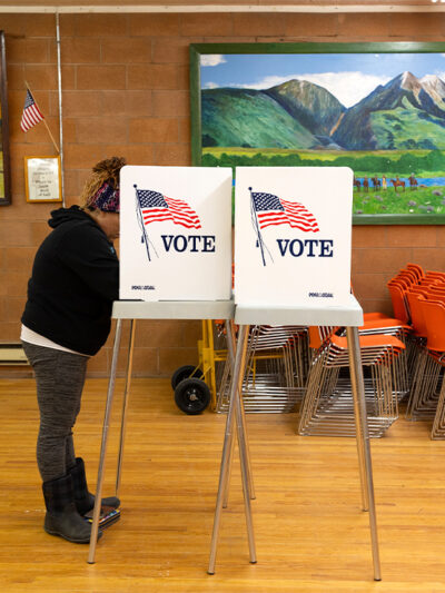 A person stands at a voting booth marked with an American flag and the word “VOTE” inside a community polling place. The room has wooden floors, orange stacked chairs, and colorful landscape paintings on the walls, with a small U.S. flag displayed nearby.