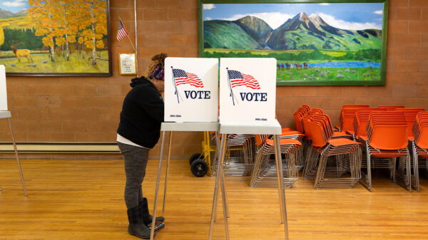 A person stands at a voting booth marked with an American flag and the word “VOTE” inside a community polling place. The room has wooden floors, orange stacked chairs, and colorful landscape paintings on the walls, with a small U.S. flag displayed nearby.