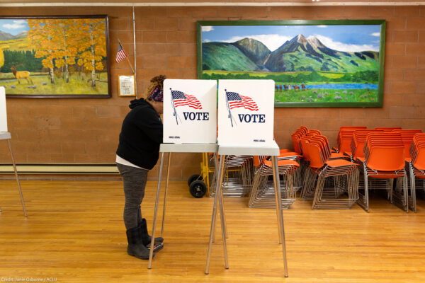 A person stands at a voting booth marked with an American flag and the word “VOTE” inside a community polling place. The room has wooden floors, orange stacked chairs, and colorful landscape paintings on the walls, with a small U.S. flag displayed nearby.