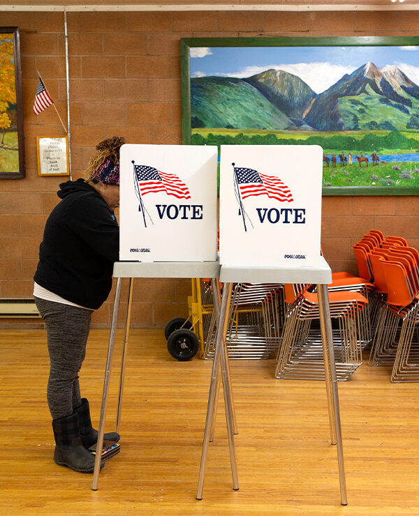 A person stands at a voting booth marked with an American flag and the word “VOTE” inside a community polling place. The room has wooden floors, orange stacked chairs, and colorful landscape paintings on the walls, with a small U.S. flag displayed nearby.