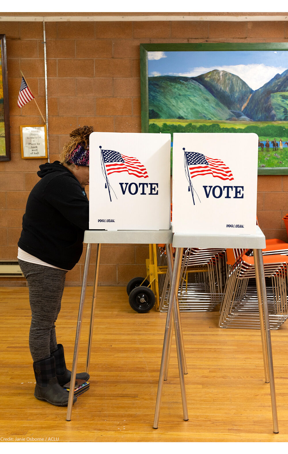 A person stands at a voting booth marked with an American flag and the word “VOTE” inside a community polling place. The room has wooden floors, orange stacked chairs, and colorful landscape paintings on the walls, with a small U.S. flag displayed nearby.