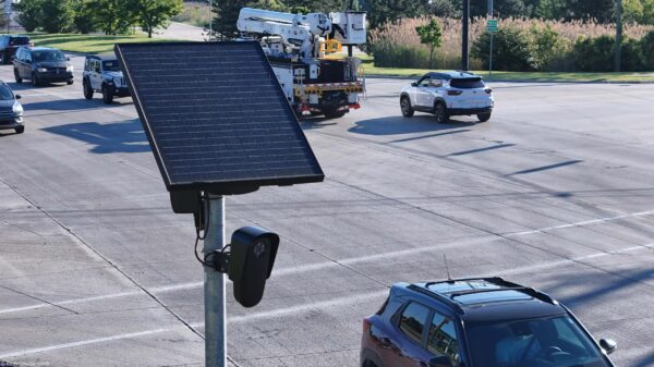 A solar-powered Flock camera is mounted on a pole beside a busy multi-lane road, with cars and a utility truck driving through the intersection in the background.