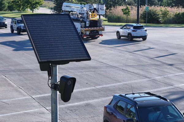 A solar-powered Flock camera is mounted on a pole beside a busy multi-lane road, with cars and a utility truck driving through the intersection in the background.