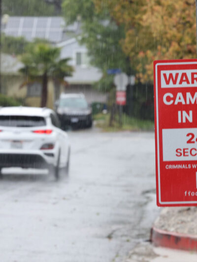 Rain falls on a residential street, where a red security sign warns "Cameras in Use 24/7 Security, Criminals Will Be Prosecuted, and flock safety." Cars drive down the wet road past houses, trees, sidewalks, and fences in the background.