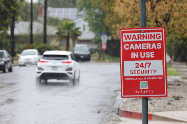 Rain falls on a residential street, where a red security sign warns "Cameras in Use 24/7 Security, Criminals Will Be Prosecuted, and flock safety." Cars drive down the wet road past houses, trees, sidewalks, and fences in the background.