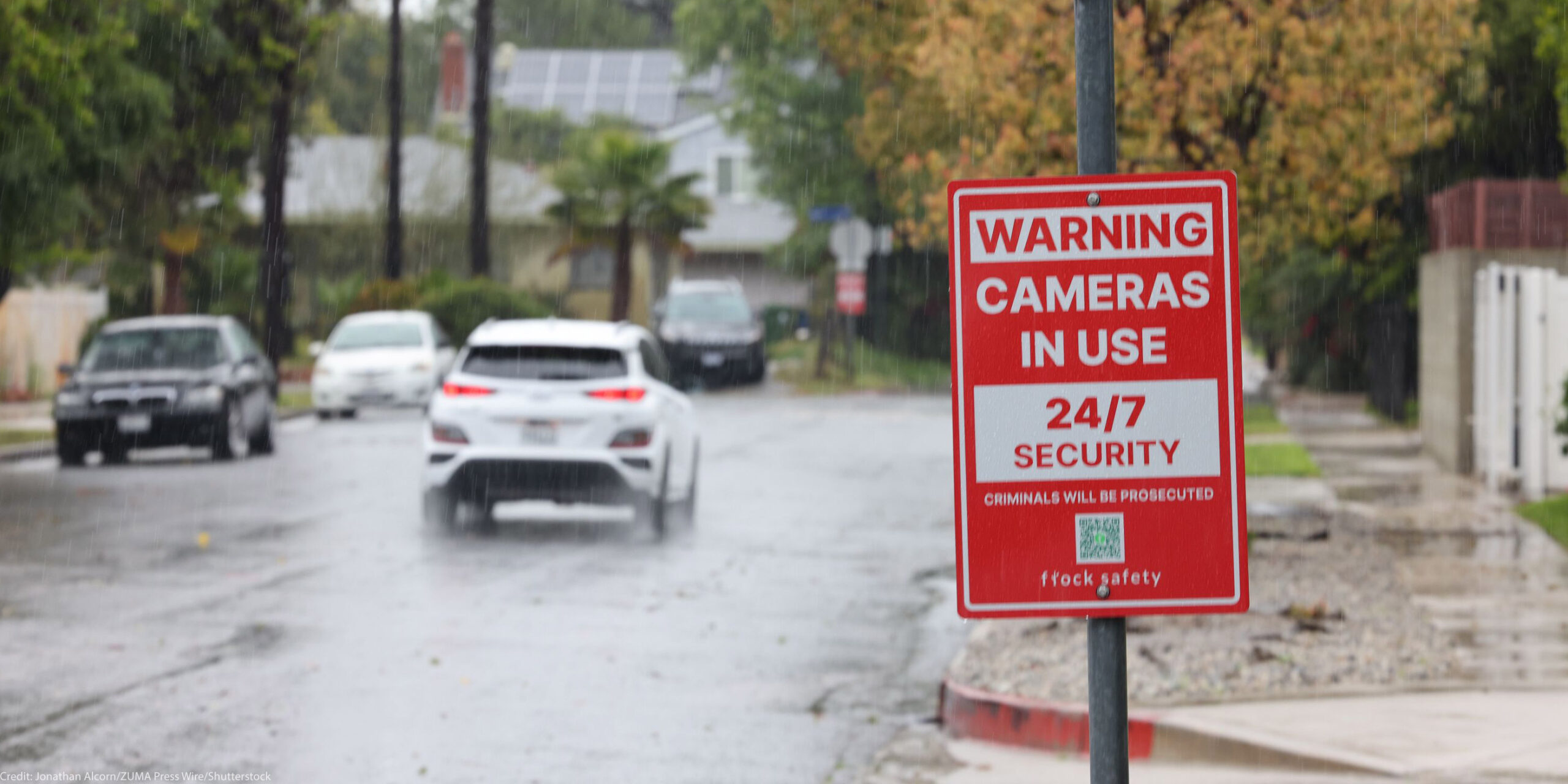 Rain falls on a residential street, where a red security sign warns "Cameras in Use 24/7 Security, Criminals Will Be Prosecuted, and flock safety." Cars drive down the wet road past houses, trees, sidewalks, and fences in the background.