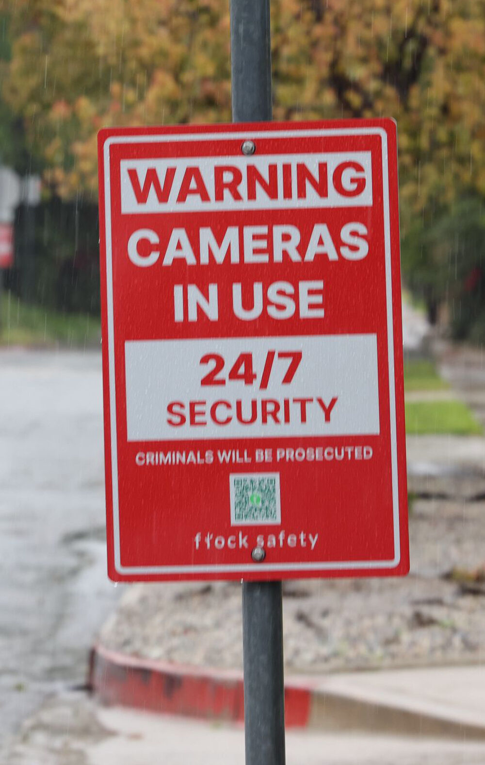 A red security sign warns "Cameras in Use 24/7 Security, Criminals Will Be Prosecuted, and flock safety." Cars drive down the wet road past houses, trees, sidewalks, and fences in the background.