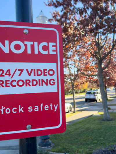 A red Flock Safety sign on a pole reads "Notice: 24/7 Video Recording" on a residential street, with parked cars, homes, trees, and a clear blue sky in the background.