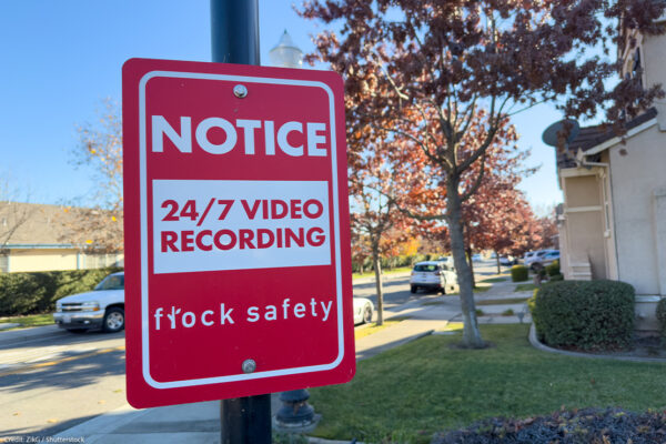 A red Flock Safety sign on a pole reads "Notice: 24/7 Video Recording" on a residential street, with parked cars, homes, trees, and a clear blue sky in the background.