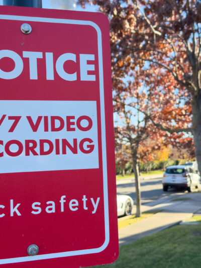 A red Flock Safety sign on a pole reads "Notice: 24/7 Video Recording" on a residential street, with parked cars, homes, trees, and a clear blue sky in the background.