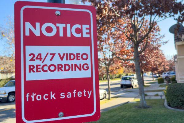 A red Flock Safety sign on a pole reads "Notice: 24/7 Video Recording" on a residential street, with parked cars, homes, trees, and a clear blue sky in the background.