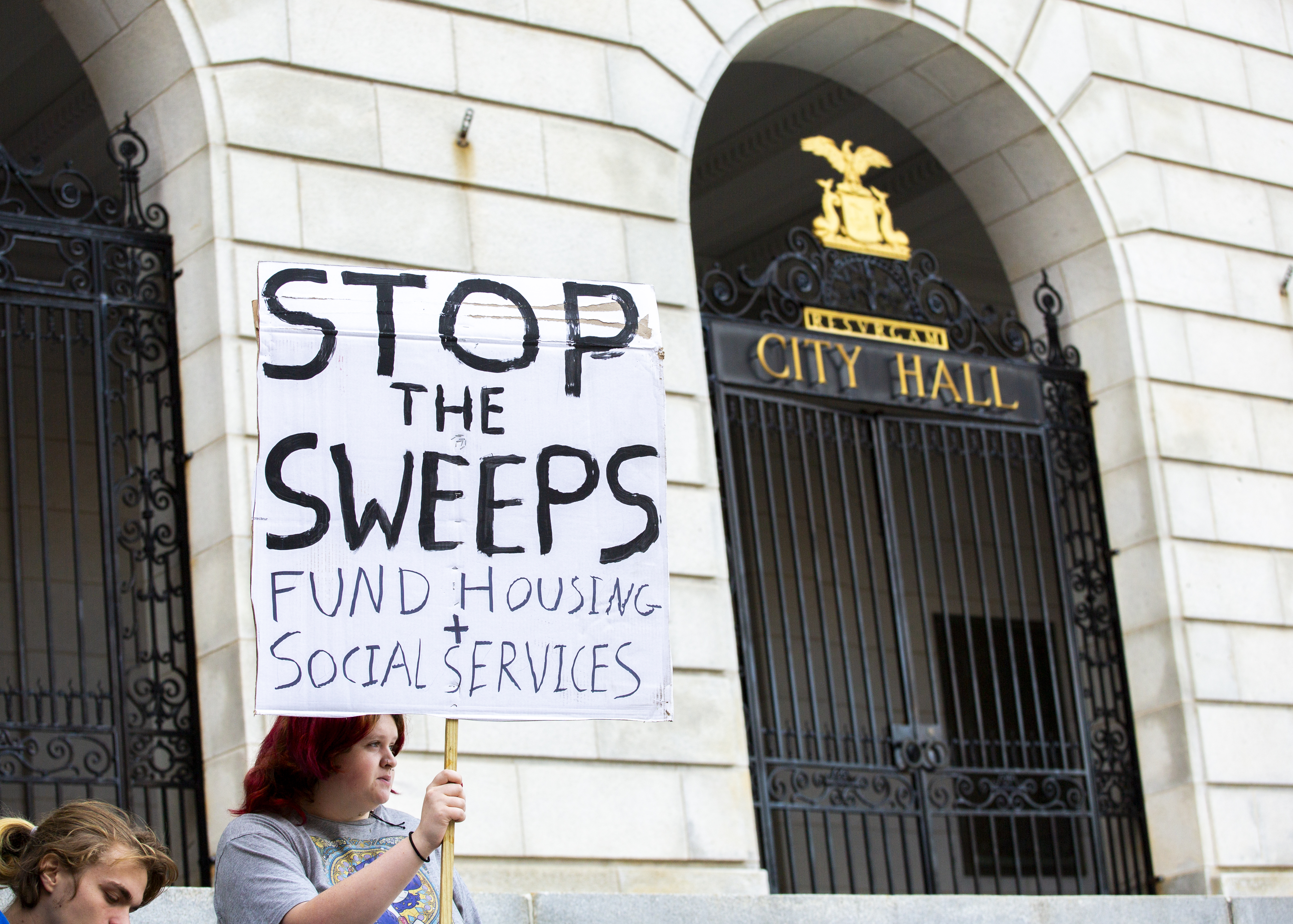 Person holding poster reading "stop the sweeps / fund housing + social services" in front of Portland City Hall.