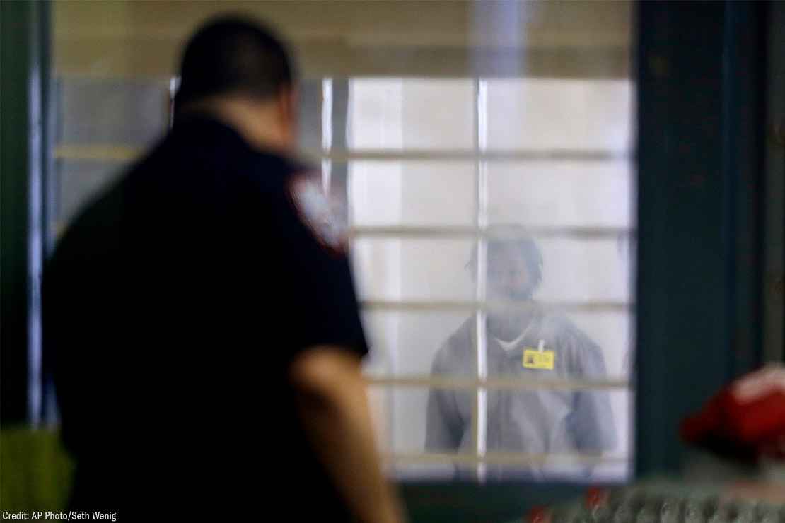 An incarcerated prison looking at a corrections officer separated by bars and a thick plastic barrier.