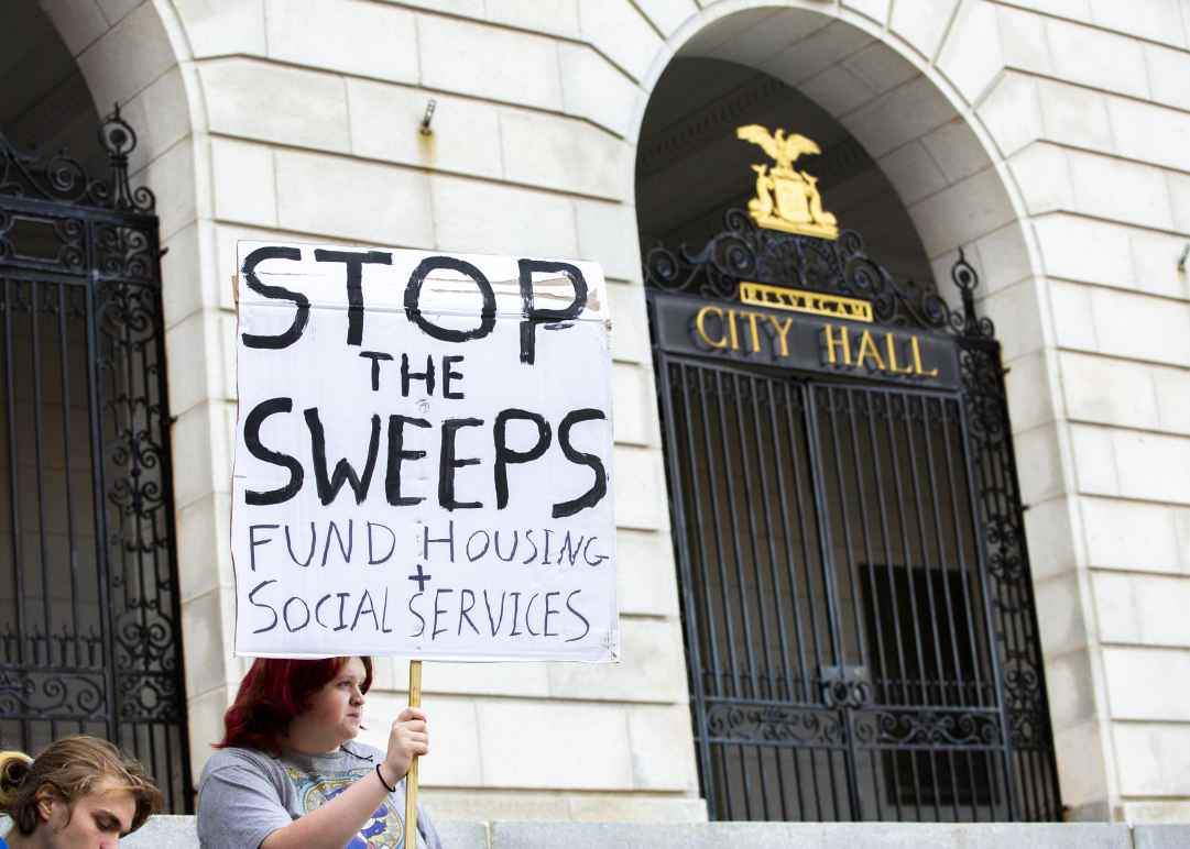 Person holding poster reading "stop the sweeps / fund housing + social services" in front of Portland City Hall.