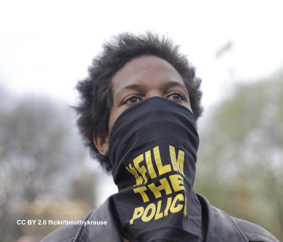 Young Black man with black bandana covering his mouth. Bandana says FILM THE POLICE in yellow letters.