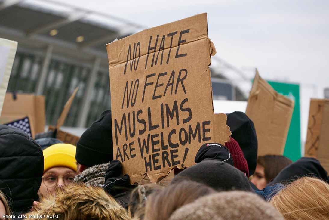 Person holding cardboard sign reading "No Hate / No Fear / Muslims Are Welcome Here!" at a protest at an airport in opposition to the Trump "Muslim Ban"
