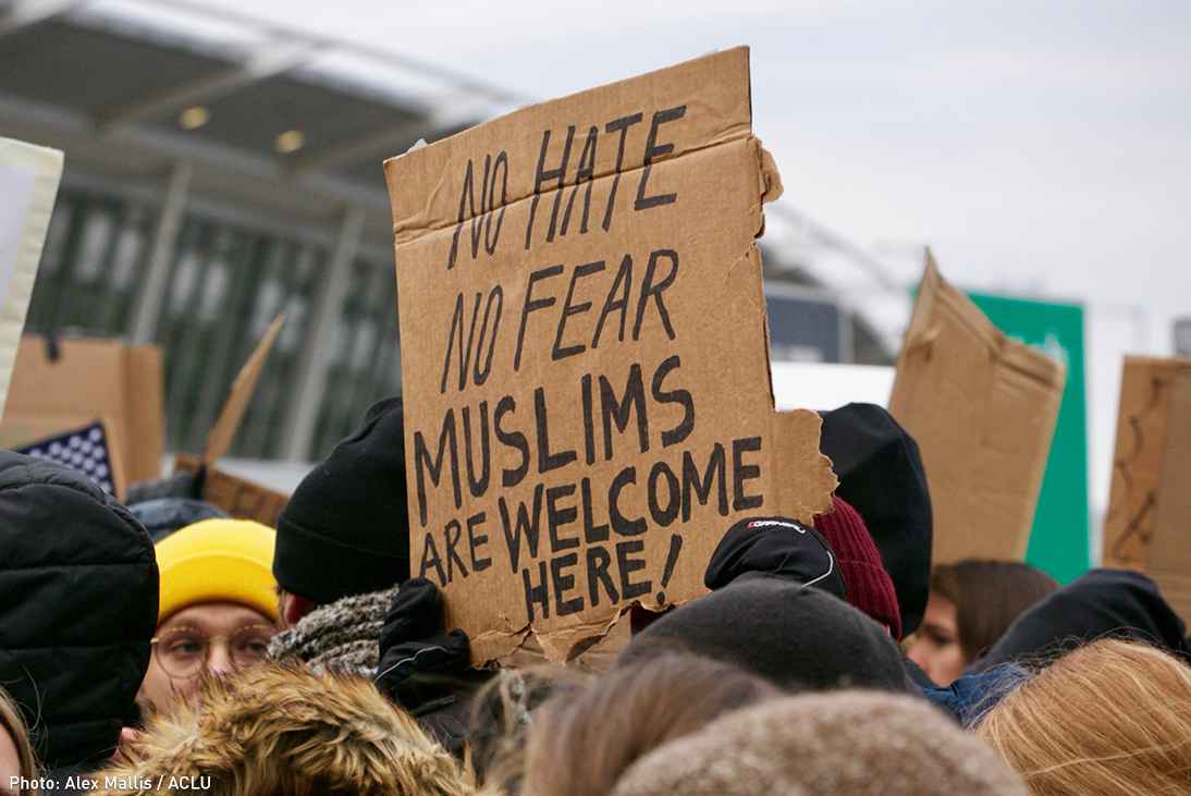 Person holding cardboard sign reading "No Hate / No Fear / Muslims Are Welcome Here!" at a protest at an airport in opposition to the Trump "Muslim Ban"