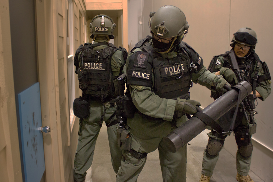 Three police officers in green uniforms preparing to knock down a door.