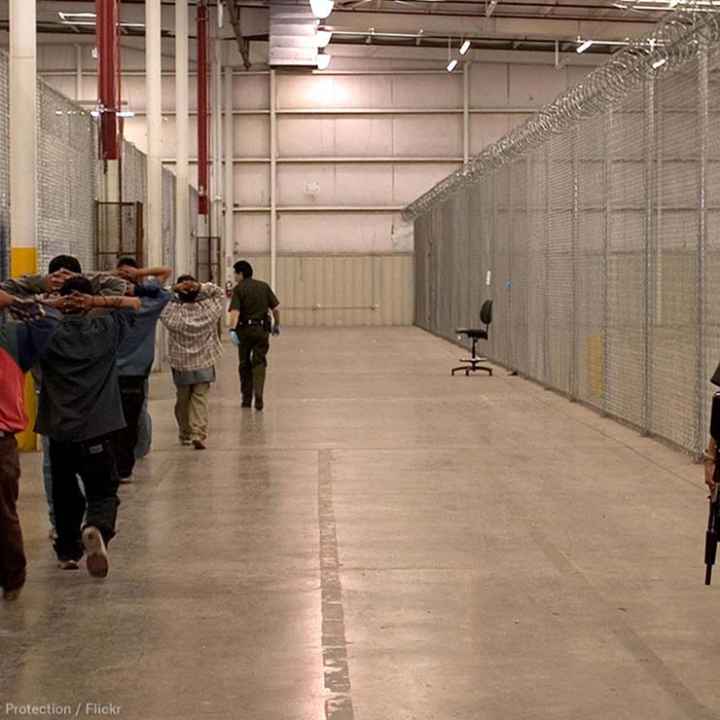 Immigrant detainees walk down a hall escorted by heavily armed CBP officers.